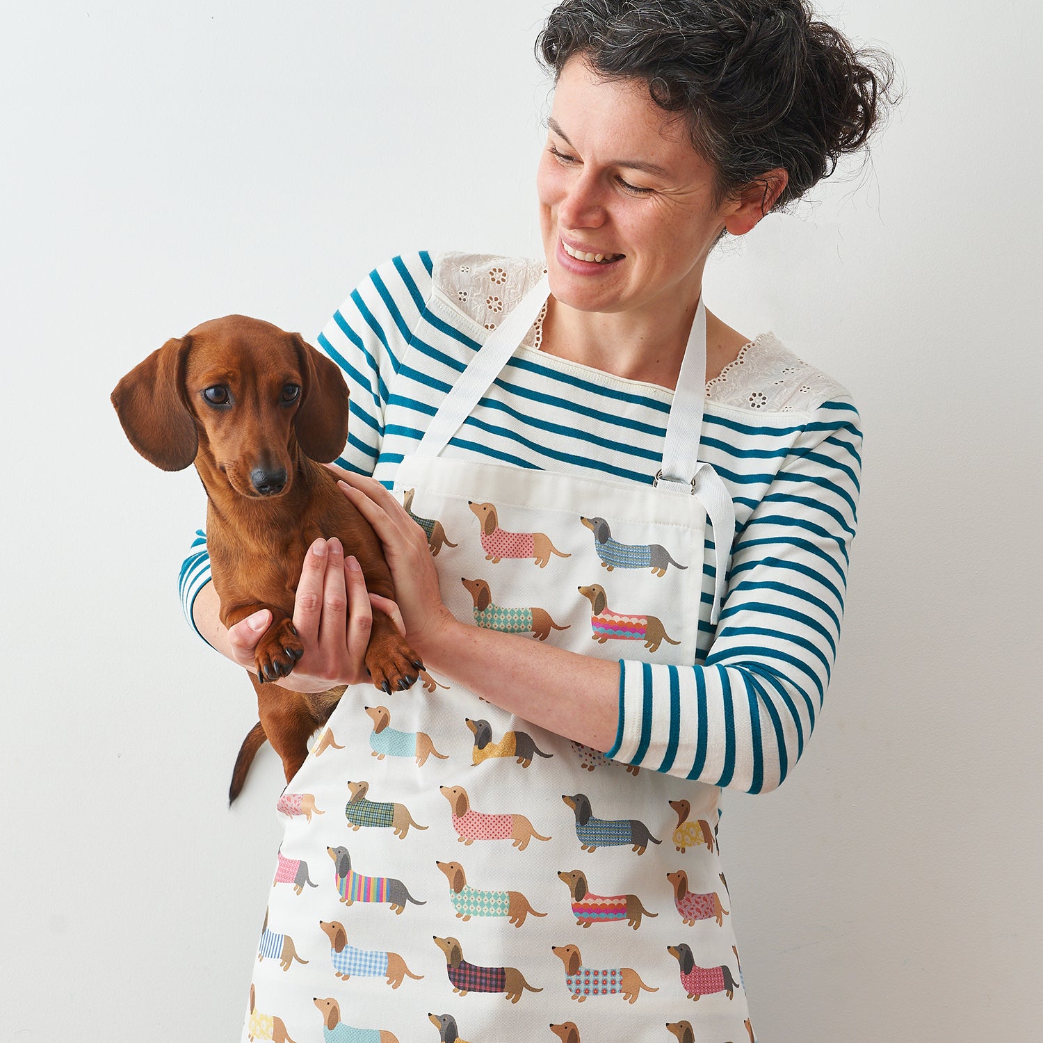 A lady holding a dachshund whilst wearing a Dashing Dachshund Apron by Mary Kilvert